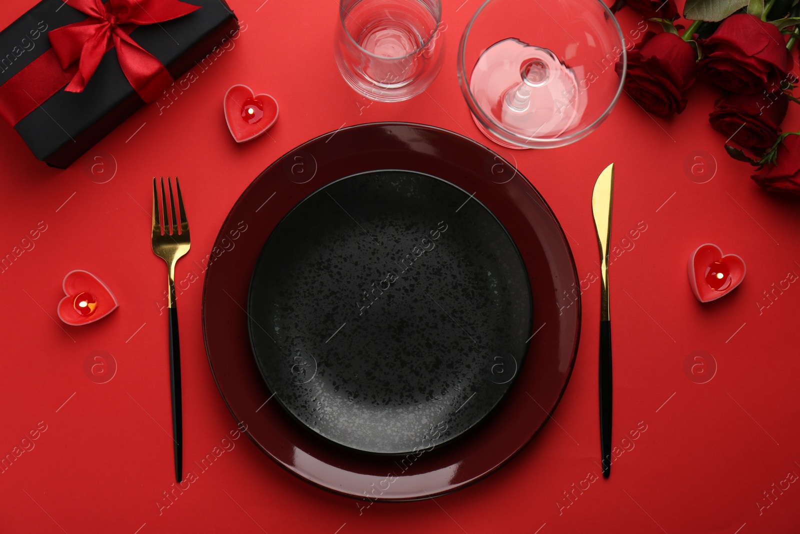 Photo of Place setting with heart shaped candles, gift box and bouquet of roses on red table, flat lay. Romantic dinner