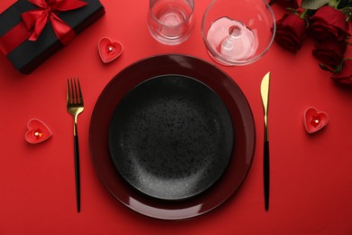 Photo of Place setting with heart shaped candles, gift box and bouquet of roses on red table, flat lay. Romantic dinner