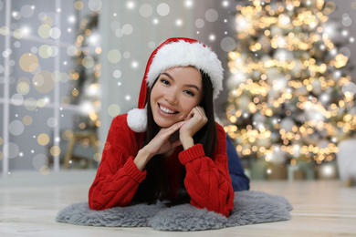 Image of Happy young woman wearing Santa hat in room with Christmas tree