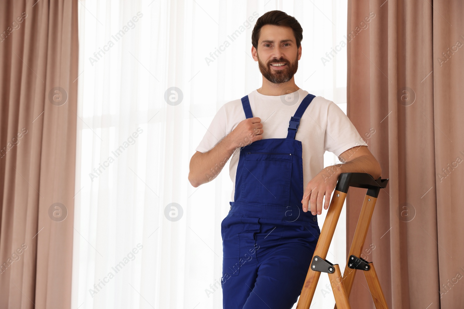 Photo of Worker in uniform standing on wooden folding ladder near window curtains indoors