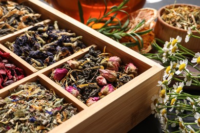 Different dry teas in wooden box and chamomile flowers on table, closeup