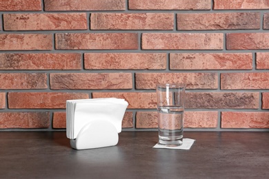 Photo of Napkin holder with paper serviettes and glass of water on table near brick wall