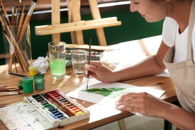 Photo of Young woman drawing leaf with watercolors at table indoors, closeup