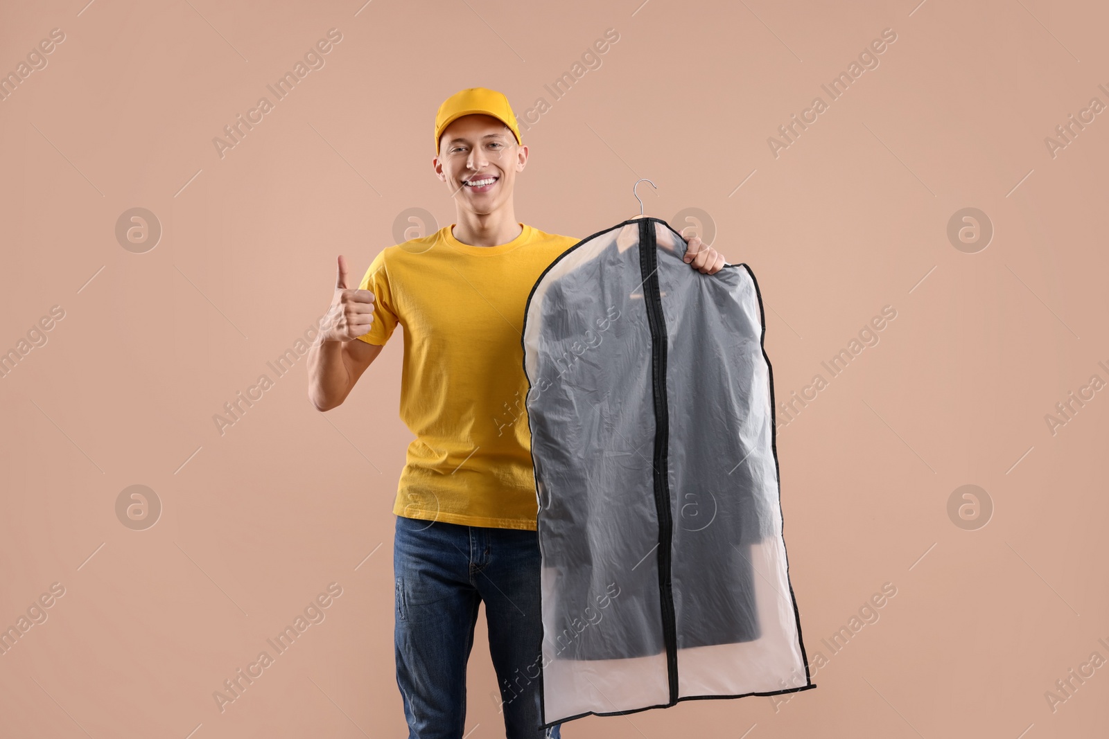 Photo of Dry-cleaning delivery. Happy courier holding garment cover with clothes and showing thumbs up on beige background