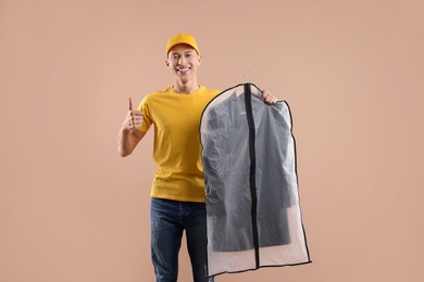 Photo of Dry-cleaning delivery. Happy courier holding garment cover with clothes and showing thumbs up on beige background