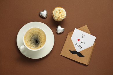 Father's day celebration. Card with phrase I Love You Daddy, cup of aromatic coffee, cupcake and heart shaped candies on brown background, flat lay