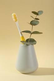 Photo of Plastic toothbrush and eucalyptus branch in holder on pale yellow background