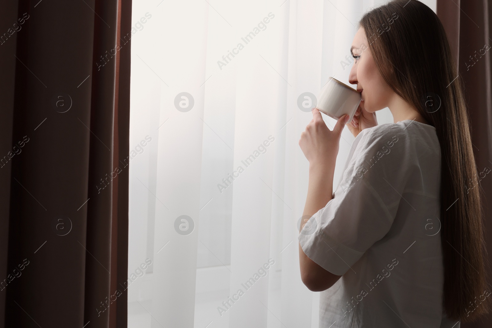 Photo of Woman holding cup of hot drink near window with stylish curtains at home. Space for text