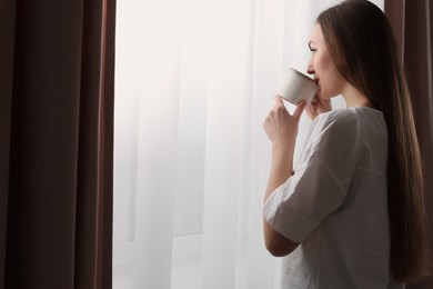 Woman holding cup of hot drink near window with stylish curtains at home. Space for text