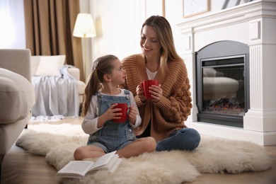 Photo of Happy woman and her daughter with cups of hot drink resting near fireplace at home