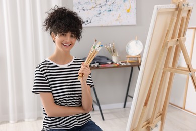 Young woman holding brushes near easel with canvas in studio