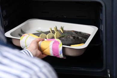 Photo of Woman putting baking tray with sea bass fish, lemon and rosemary into oven, closeup