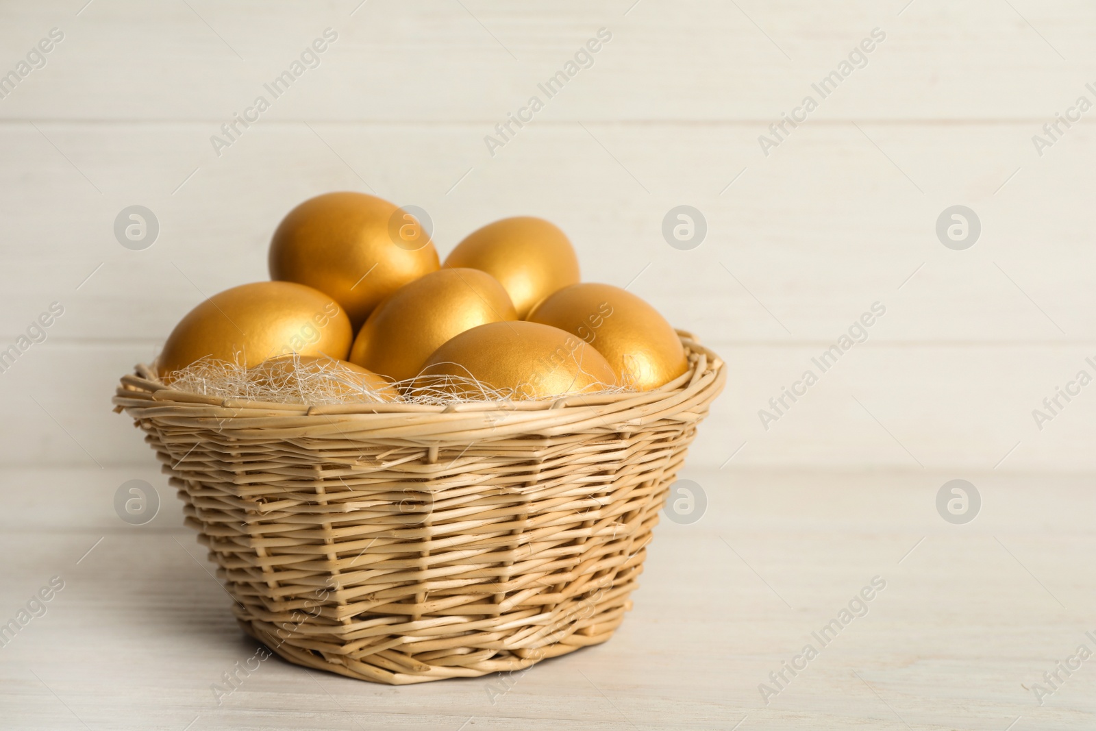 Photo of Shiny golden eggs in wicker basket on white wooden table, space for text