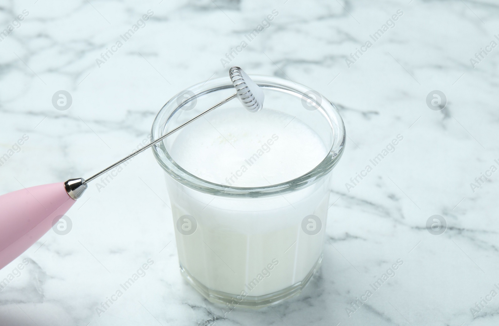 Photo of Mini mixer (milk frother) and whipped milk in glass on white marble table, closeup