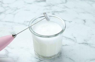 Photo of Mini mixer (milk frother) and whipped milk in glass on white marble table, closeup