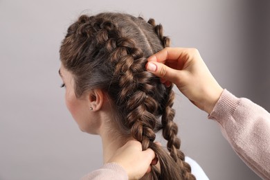 Professional stylist braiding woman's hair on grey background, closeup