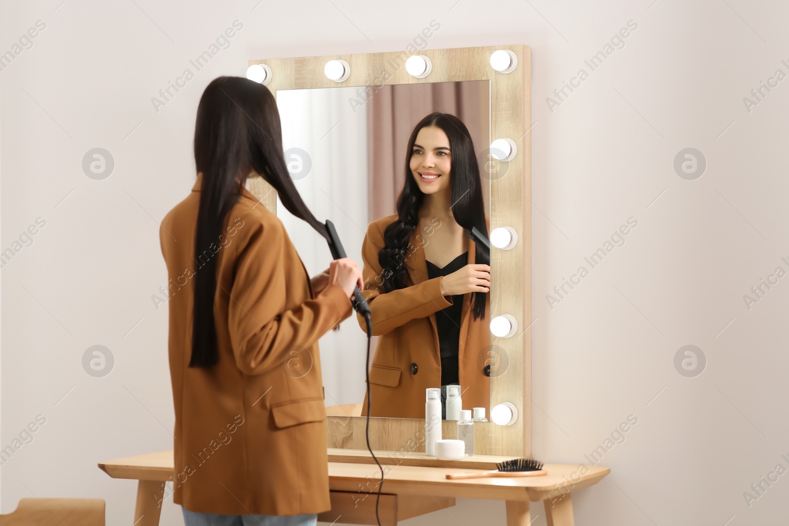 Photo of Beautiful happy woman using hair iron near mirror in room