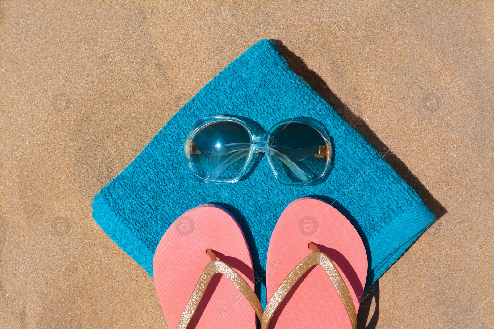 Photo of Folded soft blue beach towel with flip flops and sunglasses on sand, flat lay