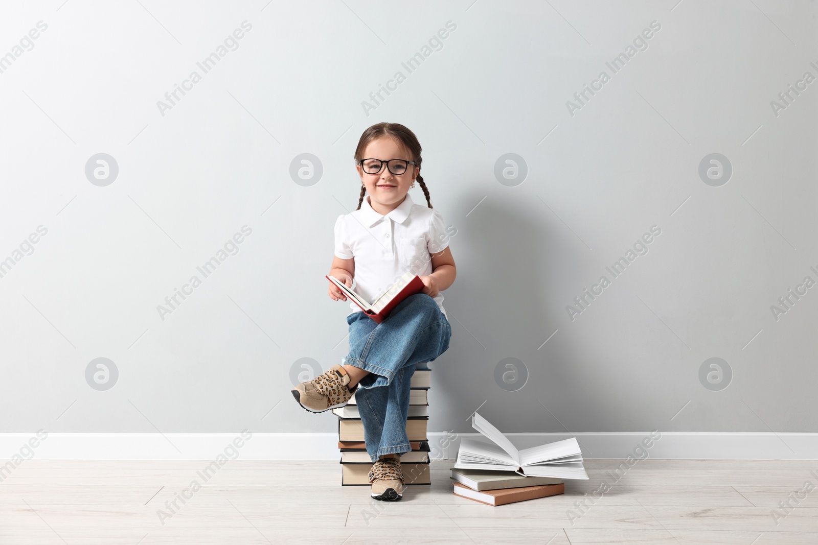 Photo of Cute little girl in glasses sitting on stack of books near light grey wall