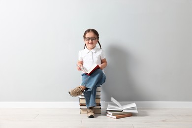 Photo of Cute little girl in glasses sitting on stack of books near light grey wall