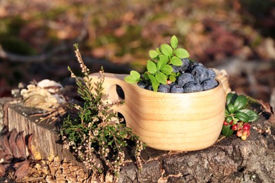 Photo of Wooden mug full of fresh ripe blueberries and lingonberries on stump outdoors