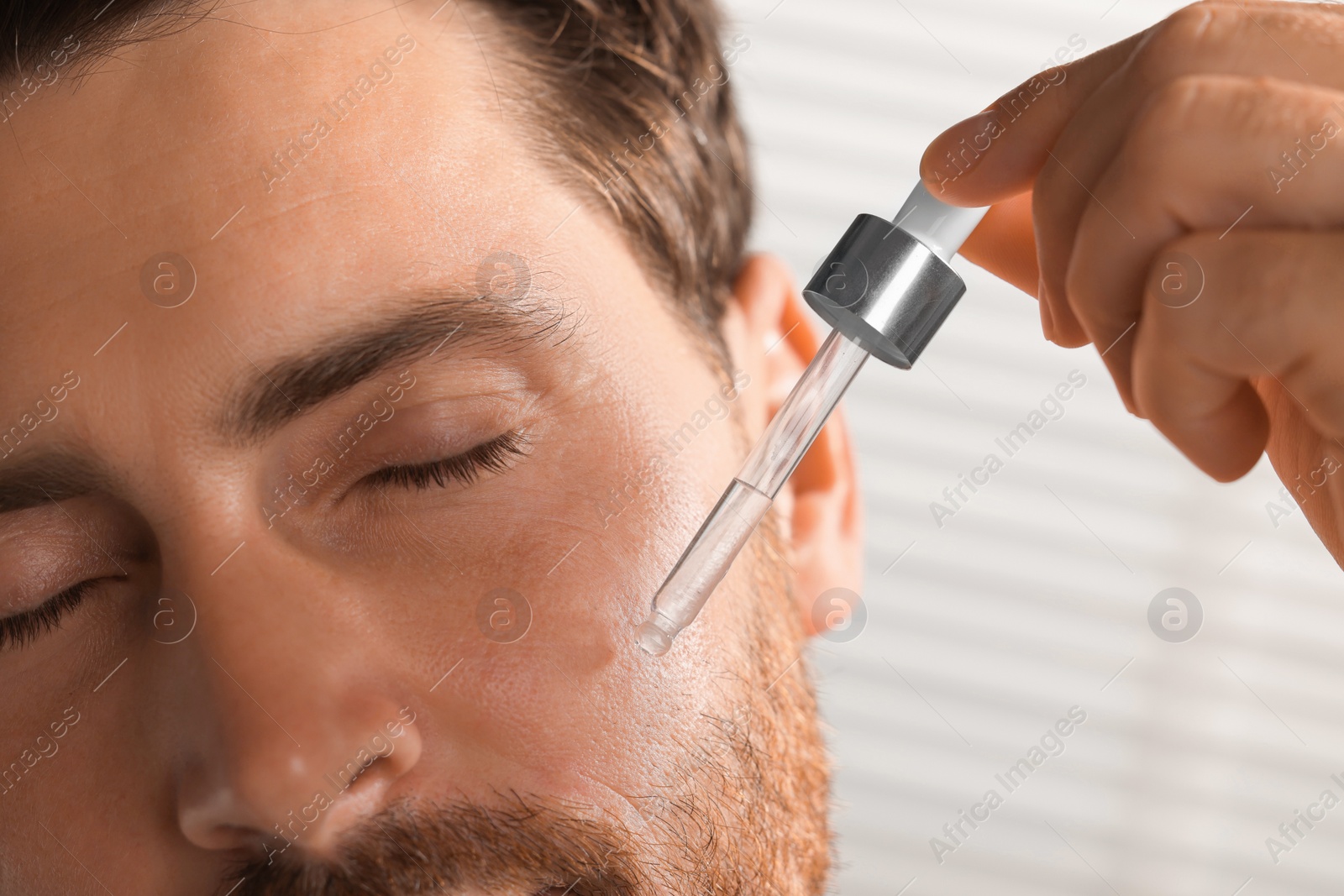 Photo of Handsome man applying cosmetic serum onto his face indoors, closeup