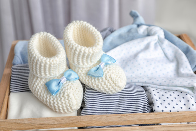 Photo of Pair of knitted booties and child's clothes in wooden crate, closeup