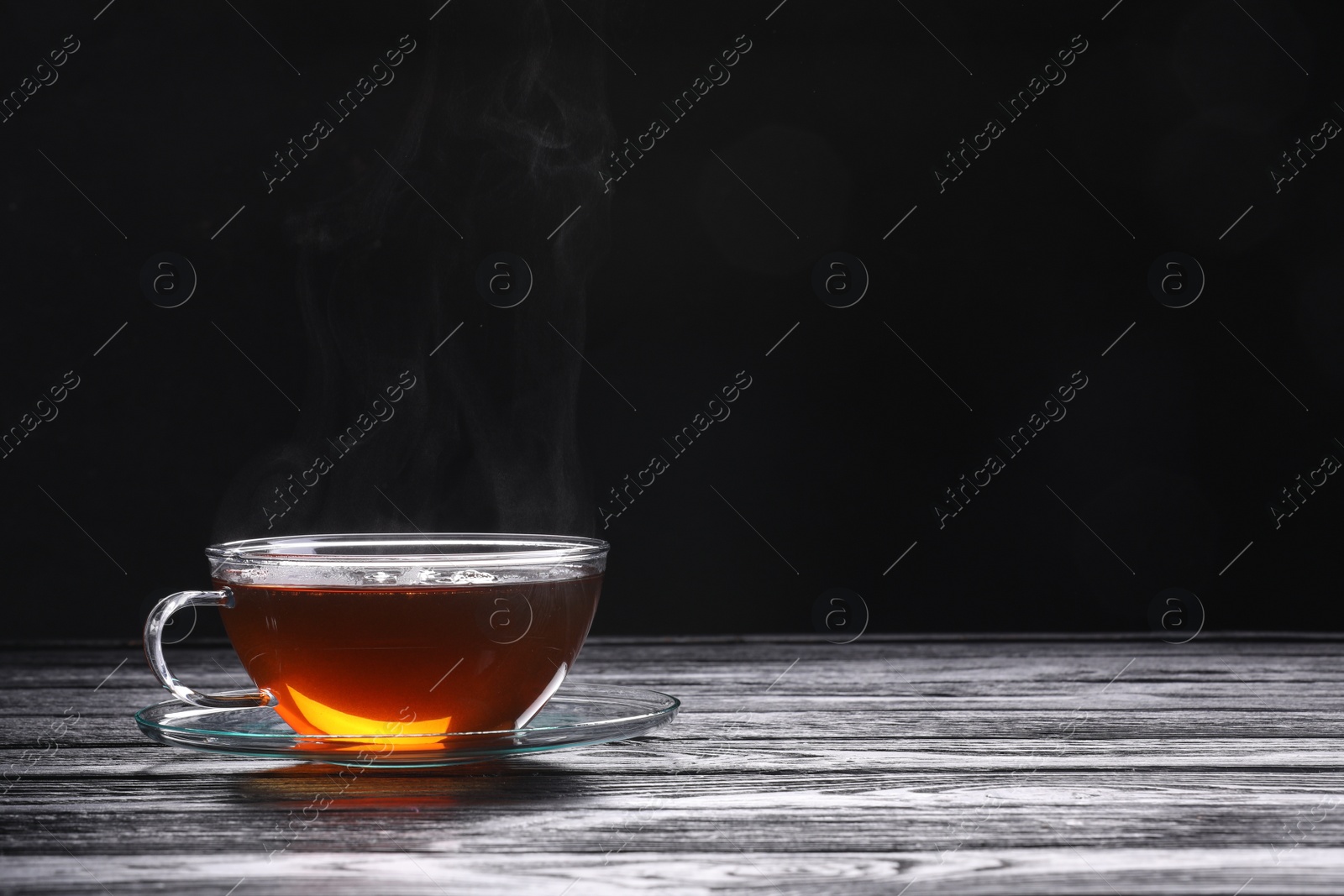 Photo of Glass cup of tea and saucer on wooden table against black background, space for text