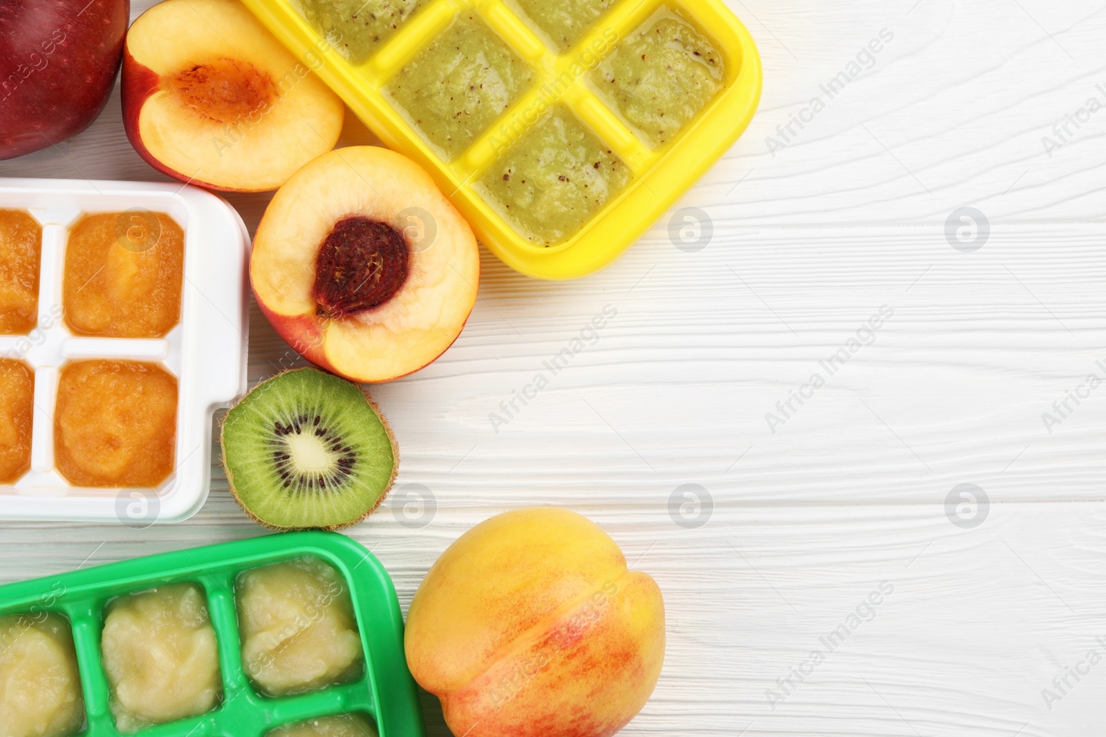 Photo of Different purees in ice cube trays ready for freezing and different fresh fruits on white wooden table, flat lay. Space for text