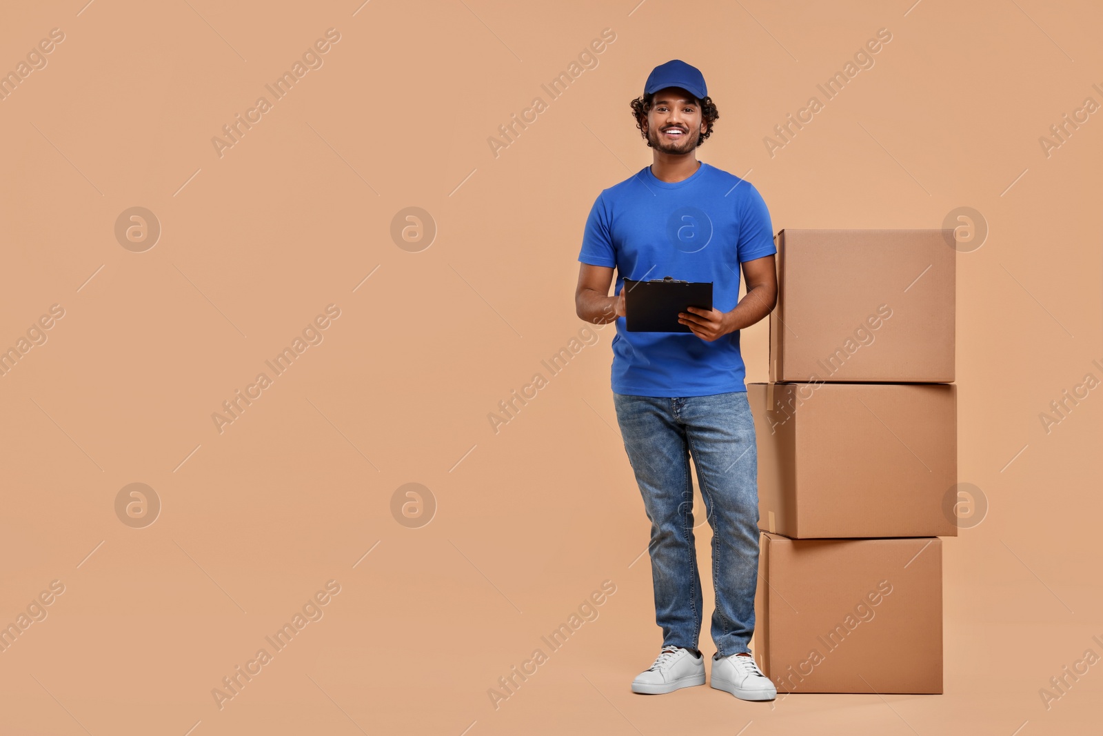 Photo of Happy courier with clipboard and stack of parcels on light brown background, space for text