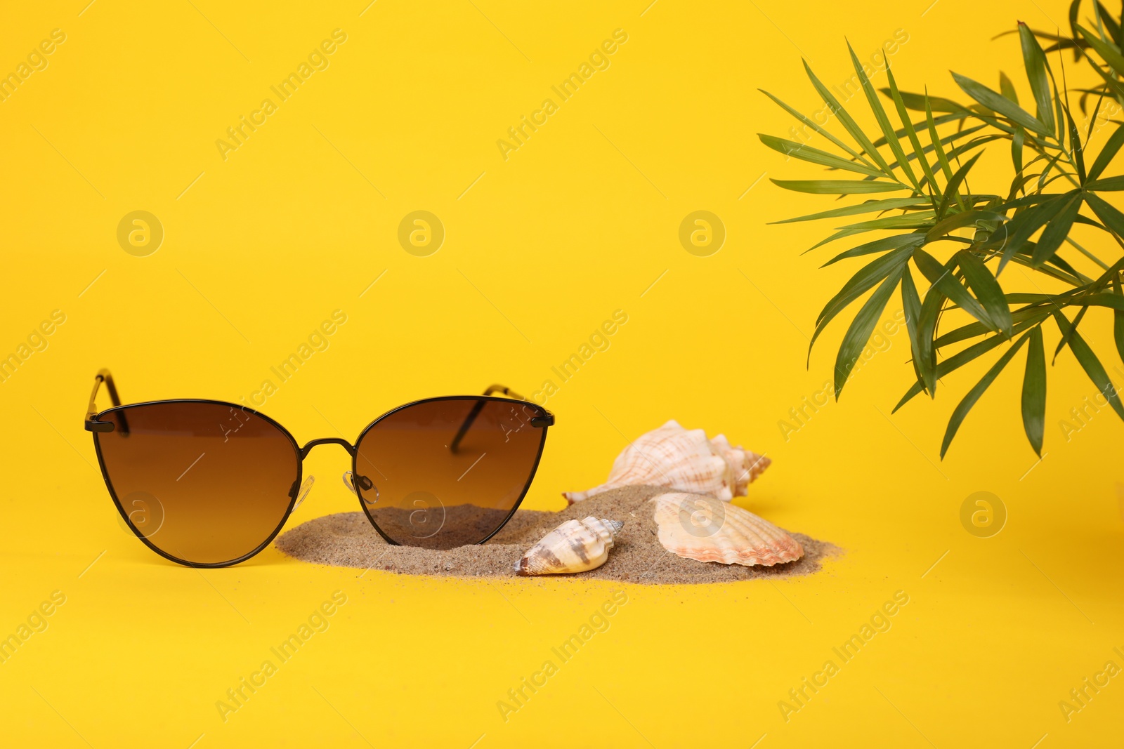 Photo of Stylish sunglasses, seashells, sand and palm leaves on yellow background