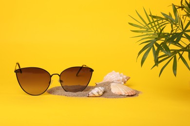 Photo of Stylish sunglasses, seashells, sand and palm leaves on yellow background