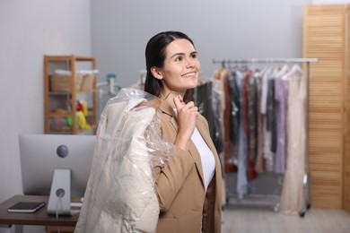 Photo of Dry-cleaning service. Happy woman holding hanger with jacket in plastic bag indoors