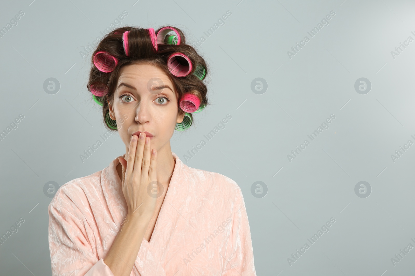 Photo of Emotional young woman in bathrobe with hair curlers on light grey background. Space for text