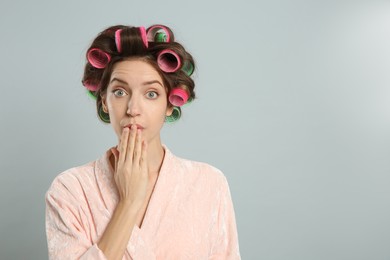 Emotional young woman in bathrobe with hair curlers on light grey background. Space for text