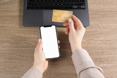 Photo of Online payment. Woman using credit card and smartphone with blank screen near laptop at wooden table, closeup