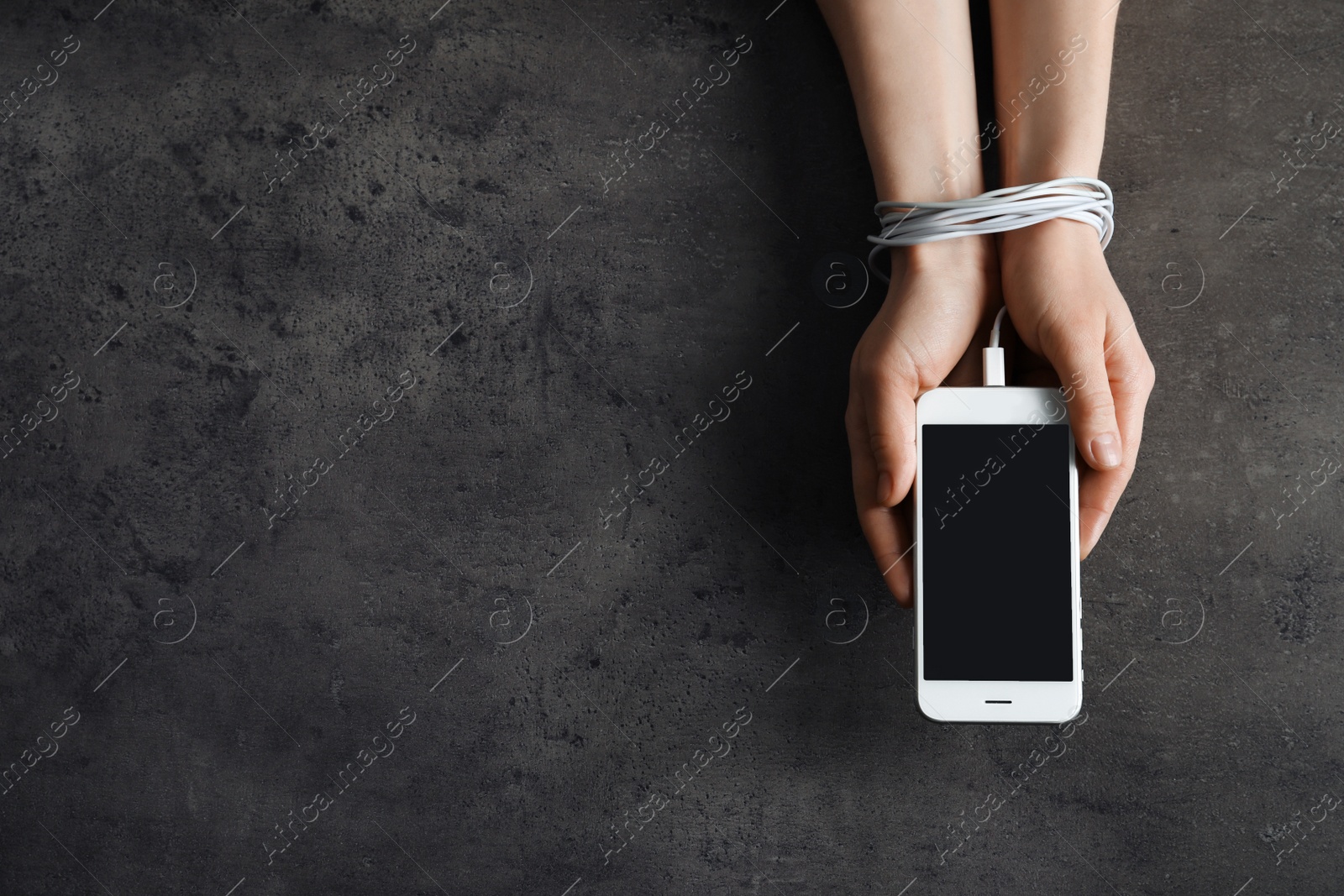 Photo of Woman holding mobile phone in hands tied with charging cable on grey background, above view. Loneliness concept