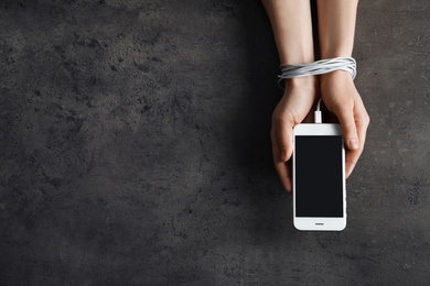 Woman holding mobile phone in hands tied with charging cable on grey background, above view. Loneliness concept