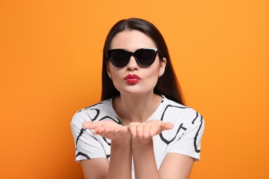 Photo of Beautiful young woman in stylish sunglasses blowing kiss on orange background