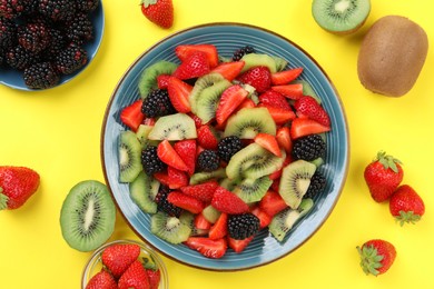 Plate of yummy fruit salad and ingredients on yellow background, flat lay
