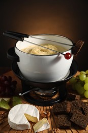 Forks with pieces of grape, bread, fondue pot with melted cheese and snacks on table, closeup