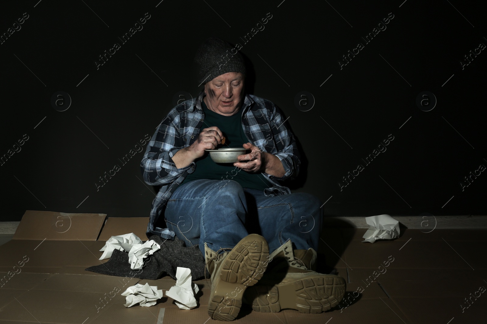 Photo of Poor senior man with bread and bowl on floor near dark wall