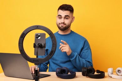 Photo of Technology blogger reviewing headphones and recording video with smartphone and ring lamp at wooden table on orange background