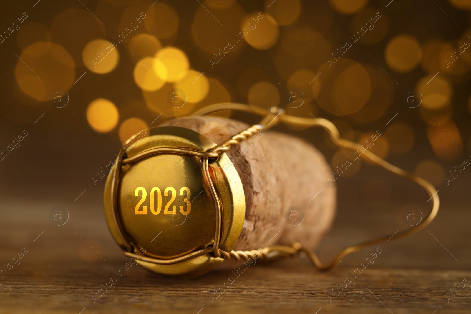 Image of Cork of sparkling wine and muselet cap with engraving 2023 on wooden table against blurred festive lights, closeup. Bokeh effect