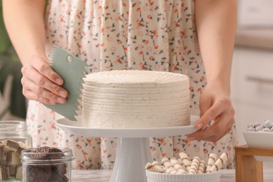 Woman using scraper to decorate cake at white table indoors, closeup
