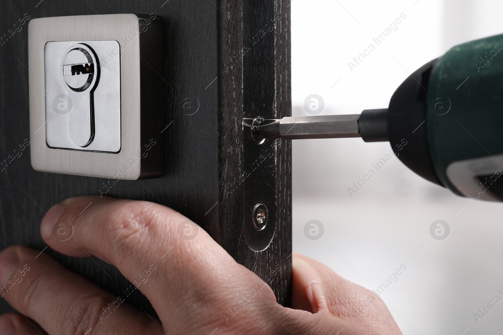Photo of Handyman with electric screwdriver repairing door lock indoors, closeup