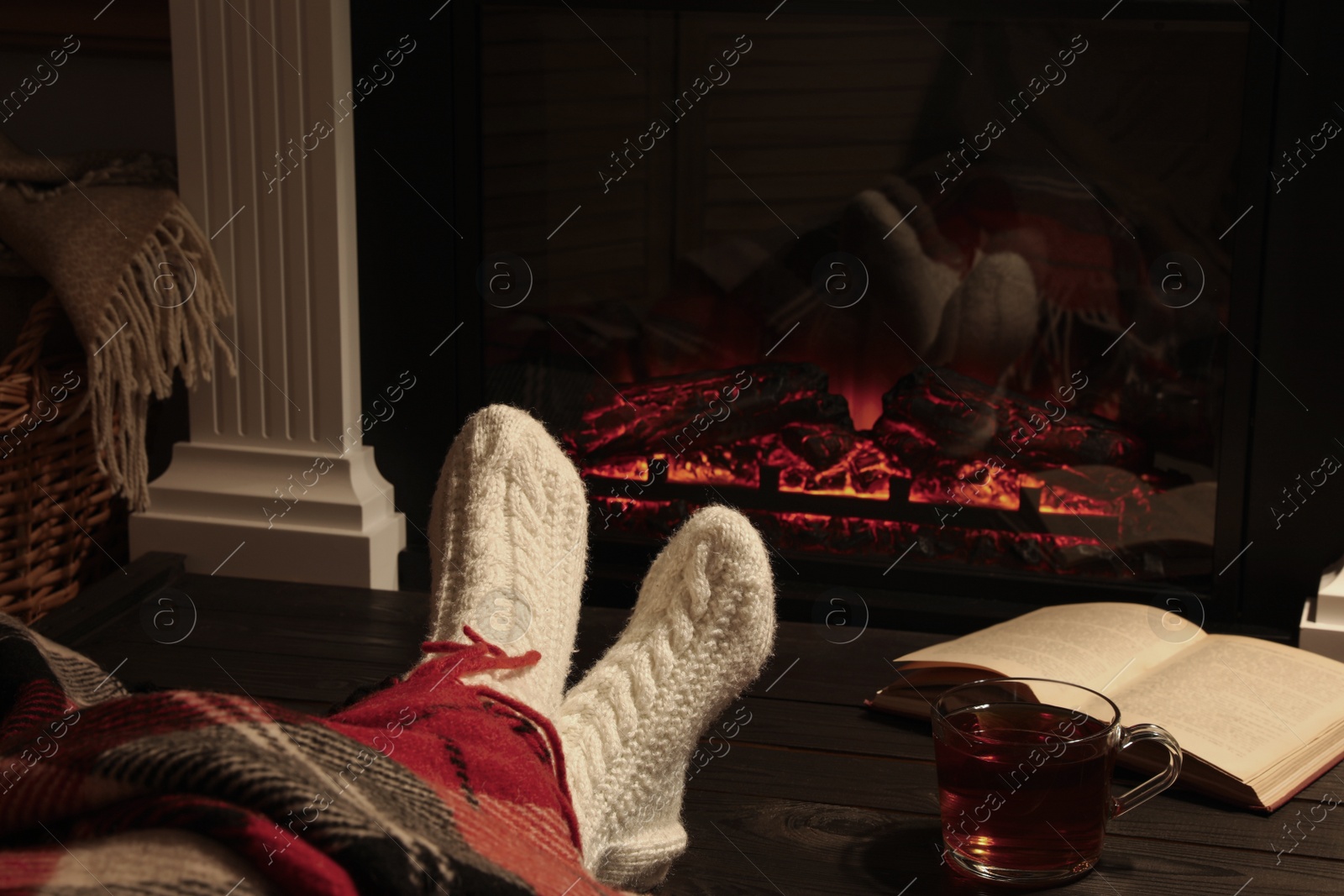 Photo of Woman with cup of tea and book resting near fireplace at home, closeup