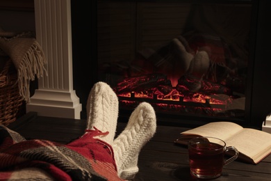 Photo of Woman with cup of tea and book resting near fireplace at home, closeup