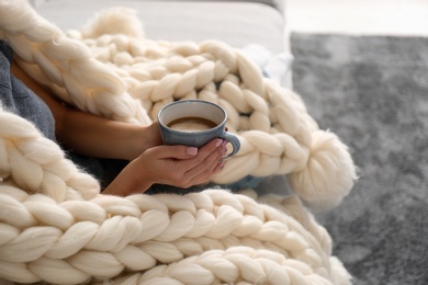 Photo of Woman covered with knitted plaid holding cup of coffee on sofa indoors, closeup