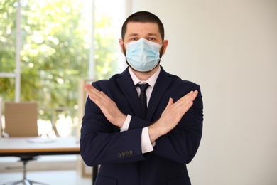 Photo of Man in protective face mask showing stop gesture in office. Prevent spreading of coronavirus